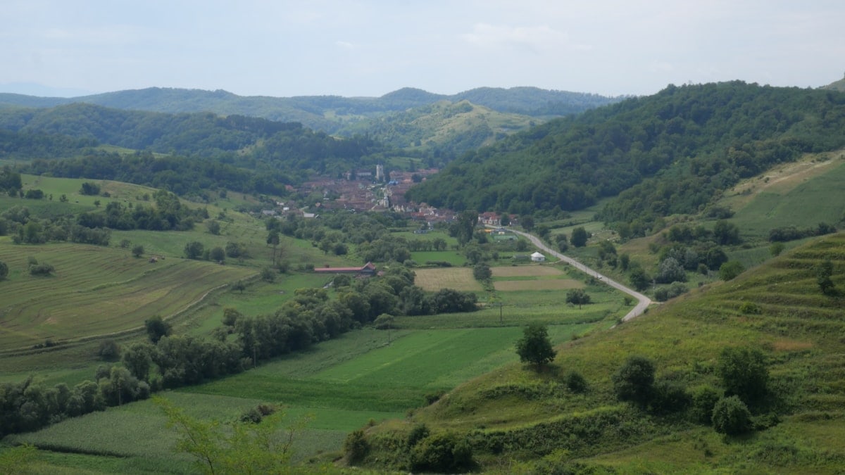A beautiful, wide-angle shot of the rural landscape in Richiș, Romania. The image features rolling green hills, a small village nestled in the valley, and a mixture of forests and fields, all under a vast, open sky.